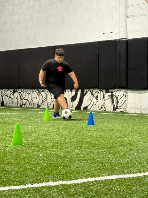 Un niño practica con el balón entre conos en una cancha de fútbol