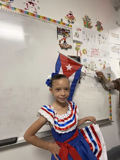 Una niña con un traje típico rojo, blanco y azul sostiene una bandera en el salón