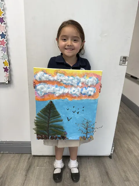 Una niña muestra una pintura de un pino y un cielo con nubes de algodón
