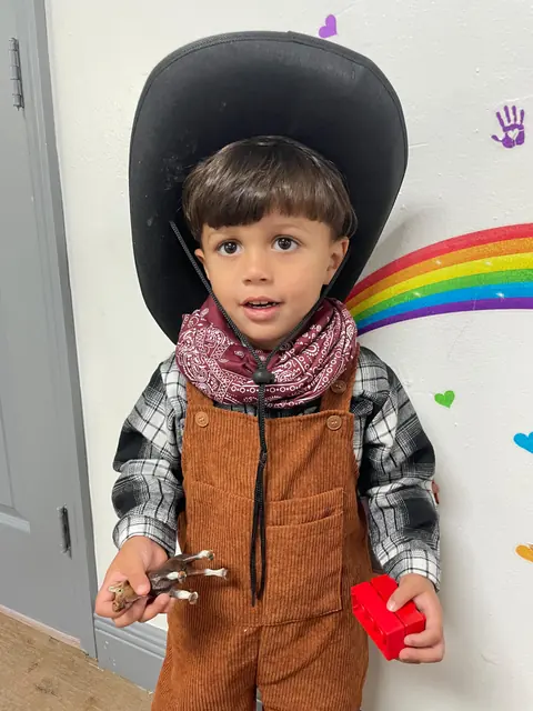 Un niño con sombrero, pañuelo y overol de vaquero sostiene un corazón rojo